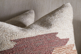 The Kabir Wool Cushion Cover - Rust from nkuku features a textured, geometric southwestern-inspired design in earthy tones. Made of a cotton and wool blend, it sits against a neutral background with another similar pillow behind it.