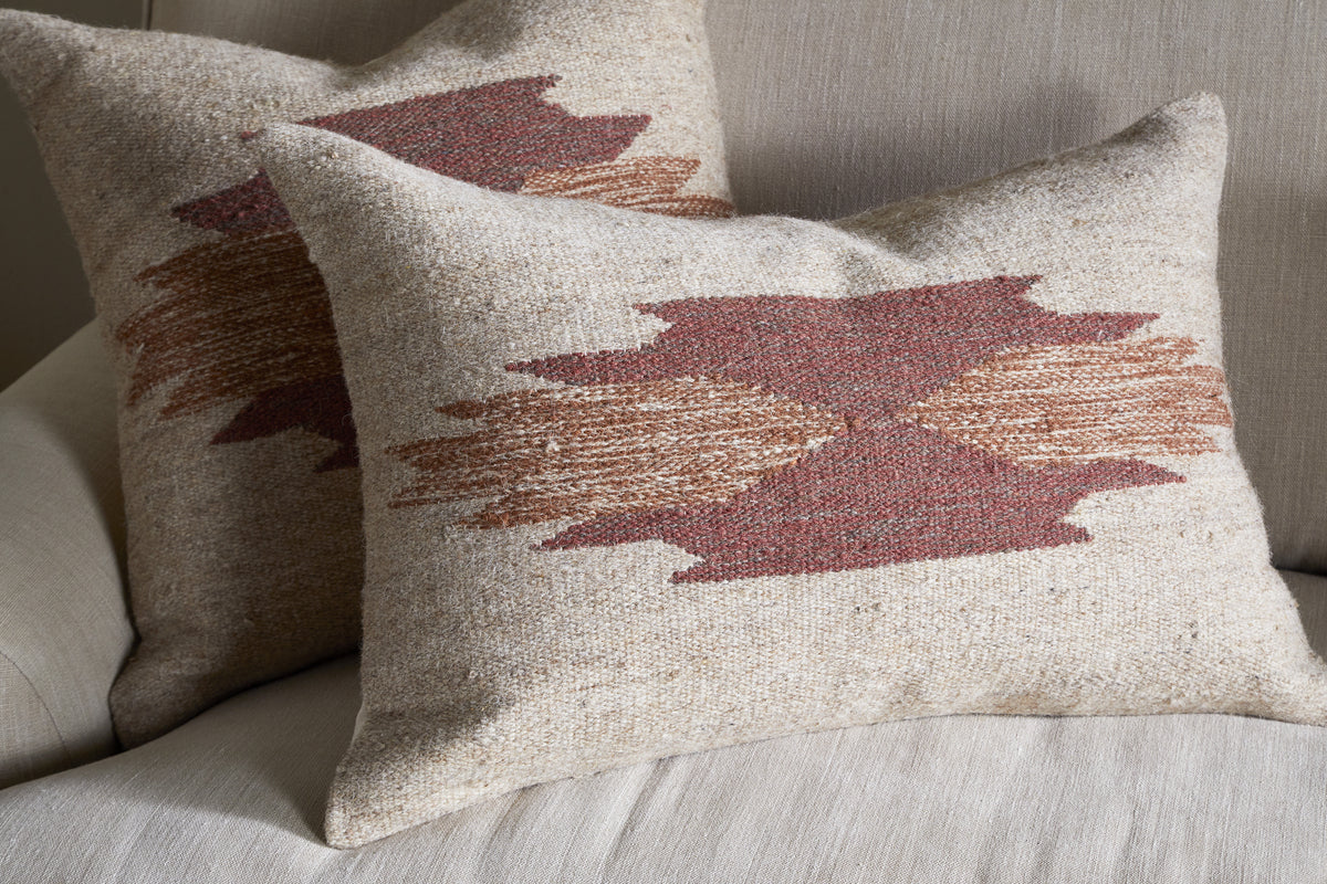 Two square throw pillows with nkuku's Kabir Wool Cushion Cover - Rust rest on a light sofa. Each features a geometric pattern in earthy red and brown tones, with overlapping zigzags forming an abstract central design.