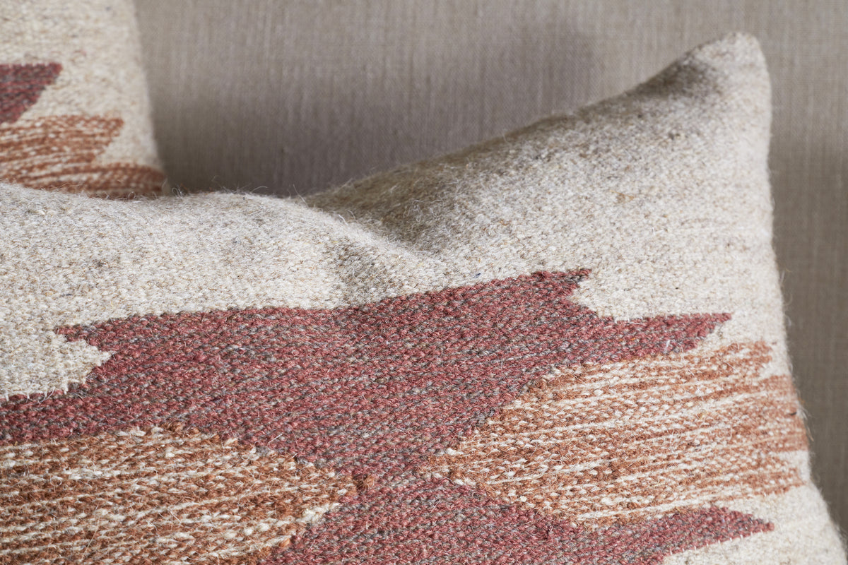 Close-up of the Kabir Wool Cushion Cover - Rust by nkuku, featuring a beige cotton and wool blend with a geometric rust and brown zigzag pattern. A second cushion in a similar design appears on the neutral fabric background.