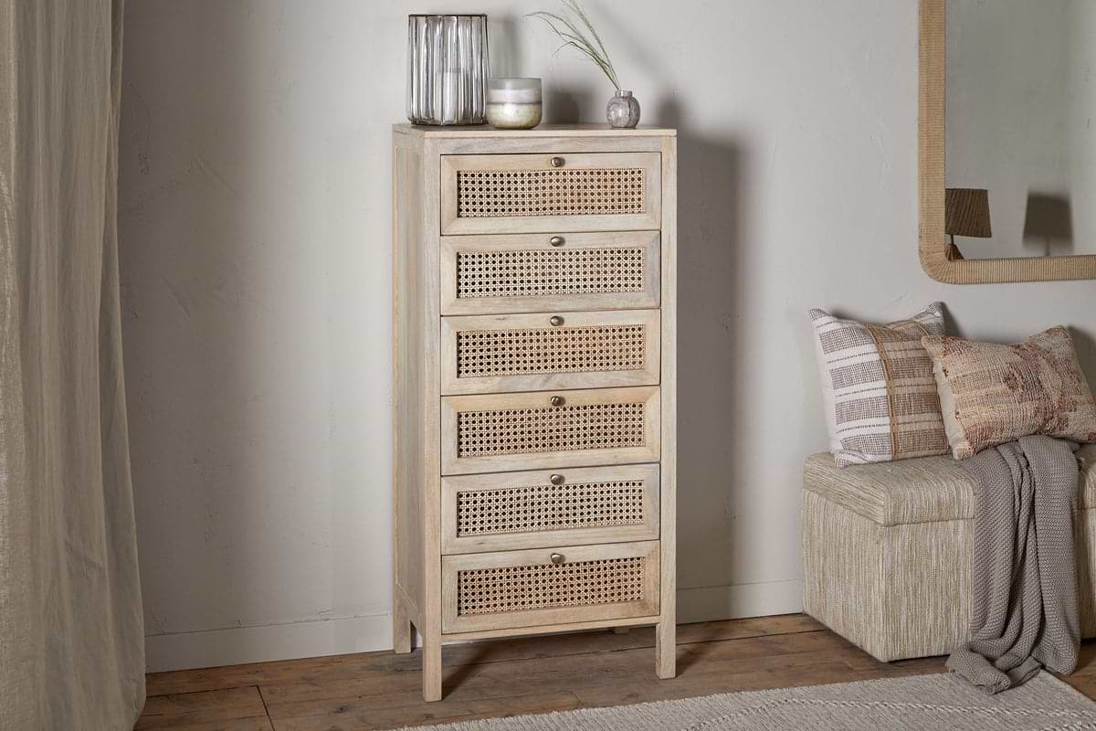 The nkuku Kanz Mango Wood Tall Boy Chest of Drawers - Natural, with six rattan cane drawers and brass knobs, stands against a white wall beside glass vases, a candle, a beige bench with textured pillows, and a mirror on wooden flooring.