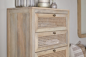 The nkuku Kanz Mango Wood Tall Boy Chest of Drawers - Natural features three cane drawers with round brass knobs. On top are two glass vases, a white candle, and a small vase with reeds. Nearby are a plaid pillow and a partial wall mirror.
