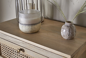 A close-up of the nkuku Kanz Mango Wood Tall Boy Chest of Drawers in Natural, with a frosted glass candle on the left and a small round marble vase with dried grass stems on the right. A tall glass candle holder appears in the background.