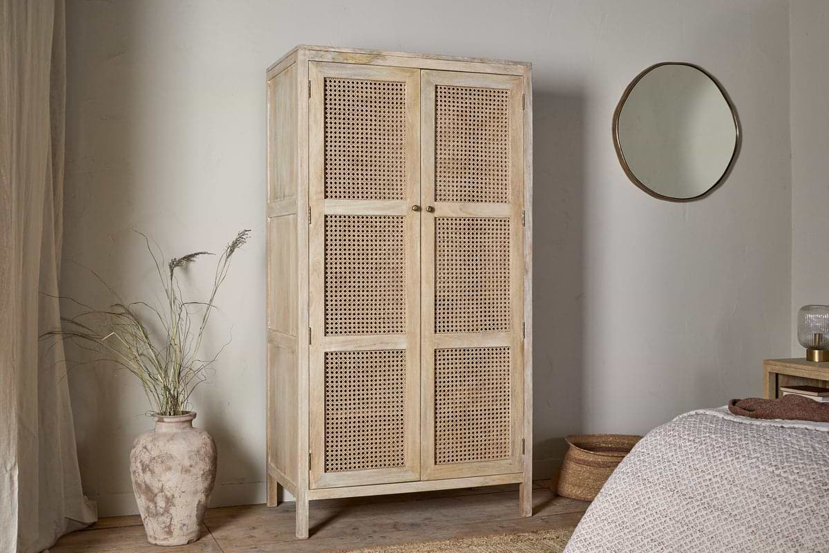 The Kanz Mango Wood Wardrobe by nkuku, featuring natural wood and woven rattan doors, stands against a white wall. A clay vase with dried grasses is on the left; to the right, a round mirror hangs above a wooden nightstand near the bed.