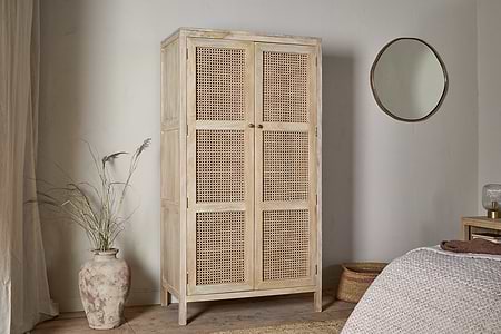 The Kanz Mango Wood Wardrobe by nkuku, featuring natural wood and woven rattan doors, stands against a white wall. A clay vase with dried grasses is on the left; to the right, a round mirror hangs above a wooden nightstand near the bed.
