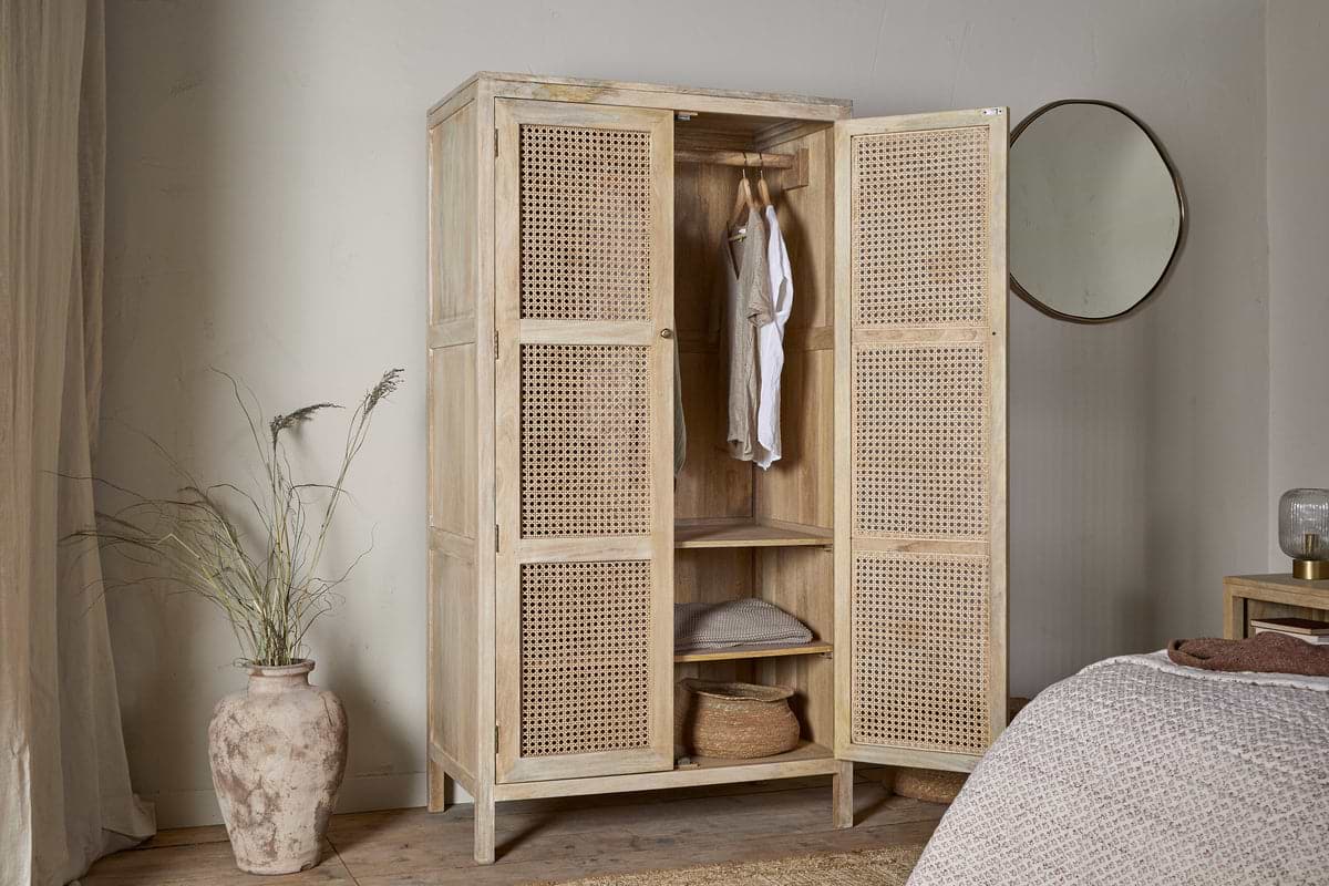 The Kanz Mango Wood Wardrobe - Natural by nkuku, a light 2-door design with woven rattan doors, stands open in a neutral bedroom. Inside are linens and baskets, with a white shirt hanging; nearby sit a round mirror, vase with grasses, and textured bedspread.