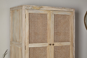 The Kanz Mango Wood Wardrobe by nkuku stands against a neutral wall, featuring double doors with woven cane panels, round metal handles, and a slightly weathered finish for a rustic look. A round mirror is partly visible to the right.