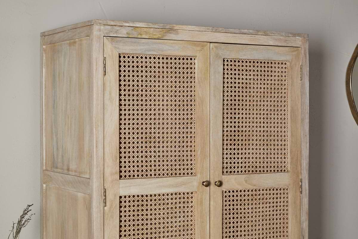 The Kanz Mango Wood Wardrobe by nkuku stands against a neutral wall, featuring double doors with woven cane panels, round metal handles, and a slightly weathered finish for a rustic look. A round mirror is partly visible to the right.