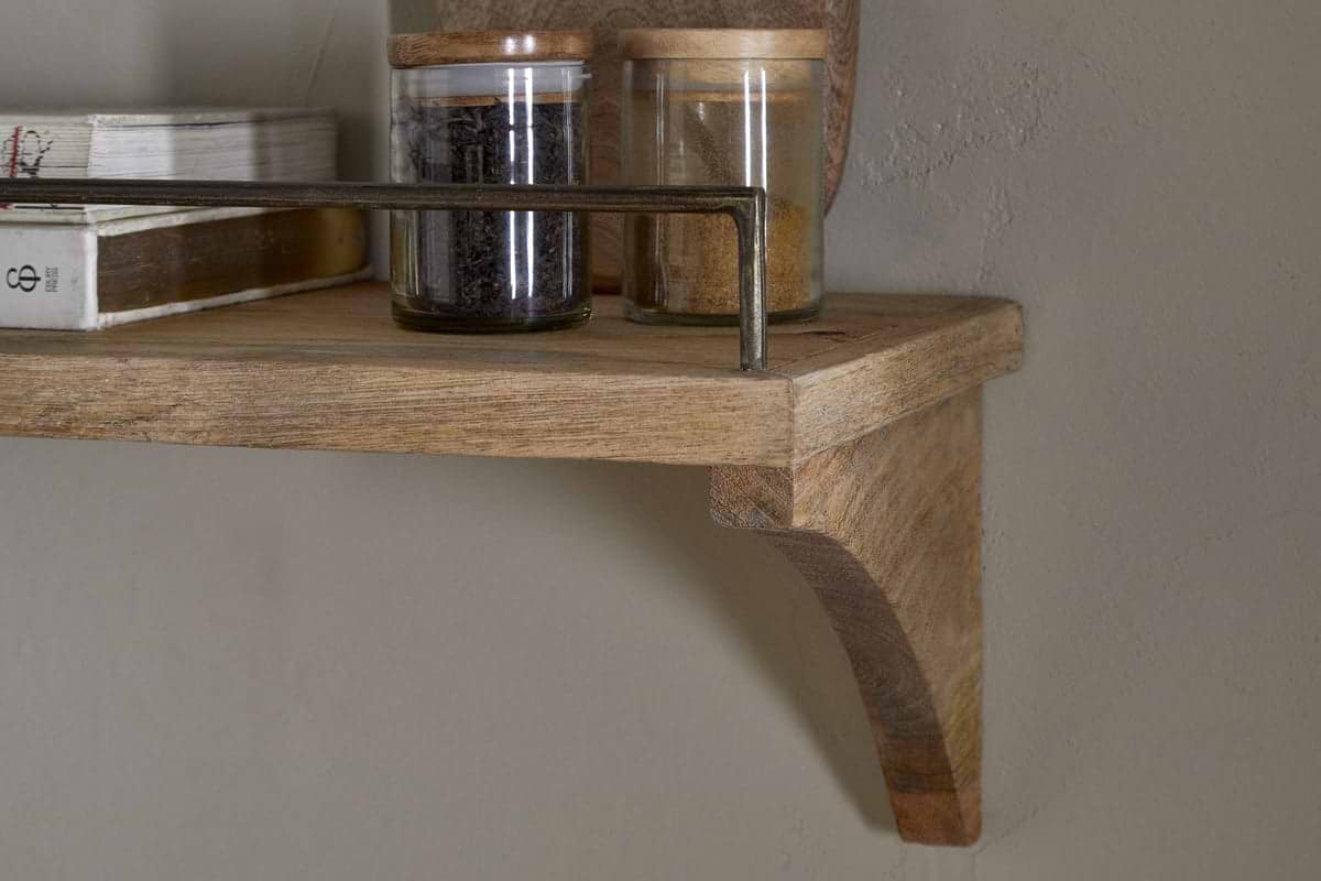 A close-up of the nkuku Meera Mango Wood & Iron Gallery Shelf - Natural, featuring a curved bracket and metal rail, holds glass jars with wooden lids filled with spices or tea leaves and books, set against a beige wall.