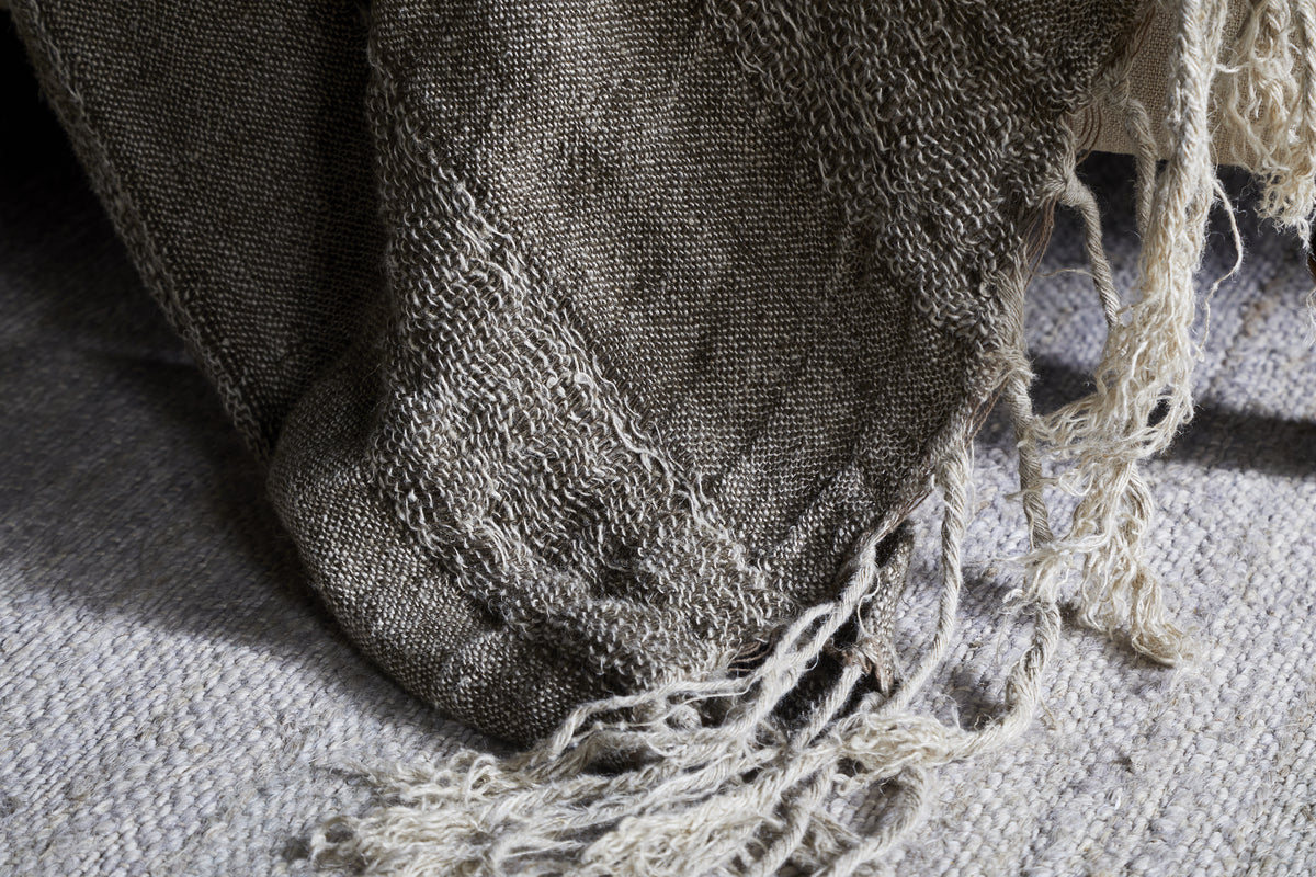 A close-up of the nkuku Munir Linen Throw in Charcoal draped over a gray rug, showcasing its slubby texture, frayed edges, and visible weave for a rustic, handmade feel.
