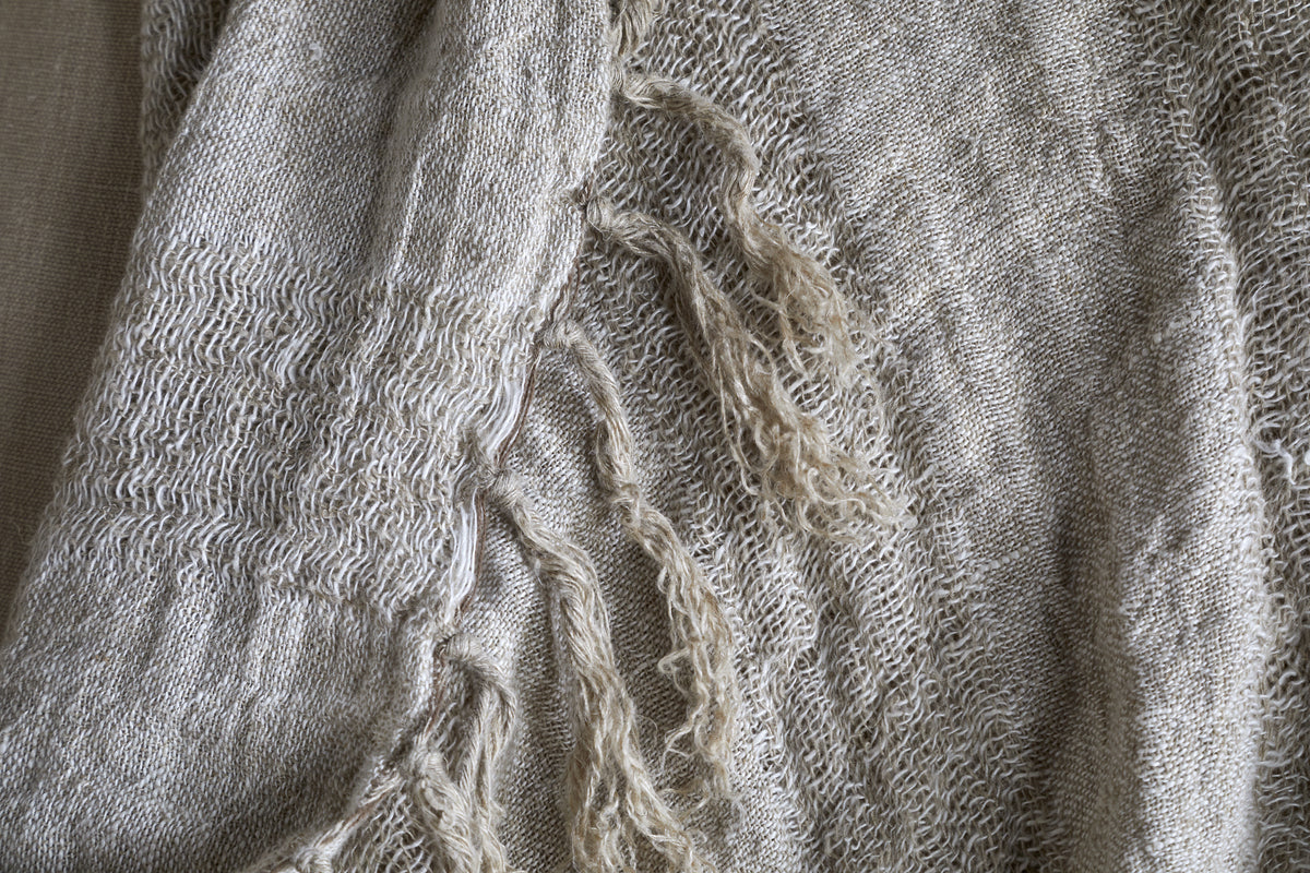 A close-up of the Munir Linen Throw - Charcoal by nkuku highlights its woven charcoal fabric with slubby texture. Frayed threads and decorative weave reveal organic, natural fibers and charming irregularities under the light.