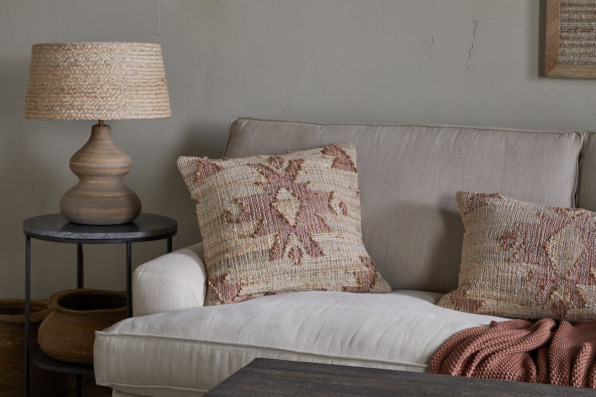 A beige sofa features two textured pillows, including the nkuku Nila Gul Cushion Cover in rust, beside a round table with a rustic lamp and woven shade. A folded terracotta throw is draped over the sofa’s arm.