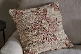 The nkuku Nila Gul Cushion Cover - Rust features a geometric, tribal-inspired pattern in earthy beige and muted rust tones, crafted from cotton and jute for a textured, rustic look that complements a light beige sofa.
