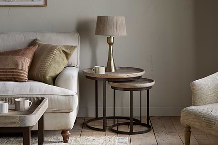 A cozy living room with a beige sofa, brown and green pillows, a gold lamp with beige shade, mugs, a small white candle, wooden floors, light walls, and nkuku’s Nisha Carved Reclaimed Wood & Iron Nesting Side Tables in Natural.