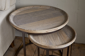 The Nisha Carved Reclaimed Wood & Iron Nesting Side Tables by nkuku, featuring a natural wood grain finish and metal legs, are arranged on a wooden floor beside a beige sofa with the smaller table partially tucked under the larger.