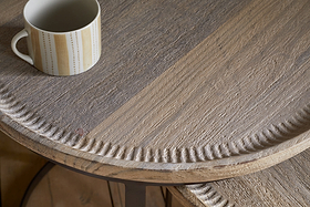 A close-up of the nkuku Nisha Carved Reclaimed Wood & Iron Nesting Side Table in Natural shows its textured light brown grain and decorative carved edge. A white mug with a vertical yellow and gray dotted line pattern sits near the upper left corner.