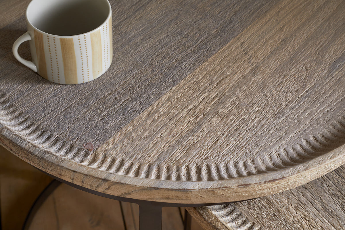 A close-up of the nkuku Nisha Carved Reclaimed Wood & Iron Nesting Side Table in Natural shows its textured light brown grain and decorative carved edge. A white mug with a vertical yellow and gray dotted line pattern sits near the upper left corner.