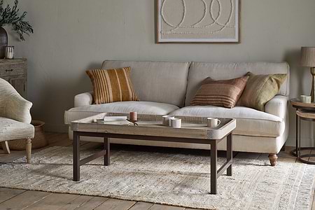 A cozy living room with a beige sofa and earth-toned pillows, light textured rug, neutral walls, framed abstract art, and a nkuku Nisha Reclaimed Wood & Iron Coffee Table - Natural topped with cups and a candle alongside a wooden side table with lamp.
