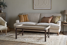 A beige sofa with earth-toned pillows rests by a light wall with abstract art. In front, nkuku's Nisha Reclaimed Wood & Iron Coffee Table - Natural stands on a patterned rug, joined by a lamp, side table, and armchair in this cozy, neutral-toned living room.