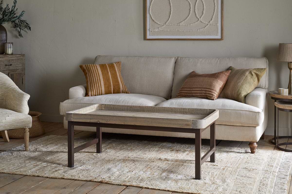 A beige sofa with earth-toned pillows rests by a light wall with abstract art. In front, nkuku's Nisha Reclaimed Wood & Iron Coffee Table - Natural stands on a patterned rug, joined by a lamp, side table, and armchair in this cozy, neutral-toned living room.