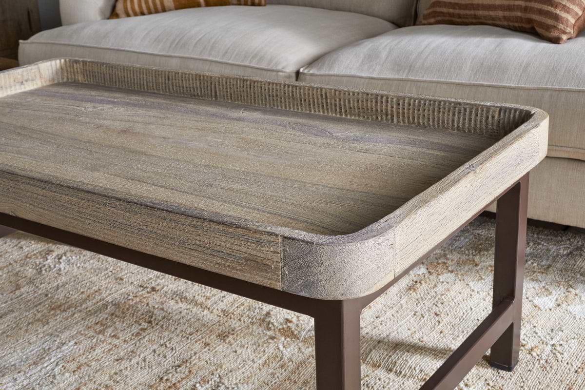 A close-up of the nkuku Nisha Reclaimed Wood & Iron Coffee Table – Natural, featuring raised edges, a rustic finish, and dark metal legs. The table sits on a light textured rug before a beige sofa with striped cushions.