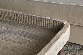 Close-up of the nkuku Nisha Reclaimed Wood & Iron Coffee Table - Natural, featuring a raised textured edge and weathered finish with visible wood grain. A beige upholstered sofa and part of a patterned rug appear in the background.