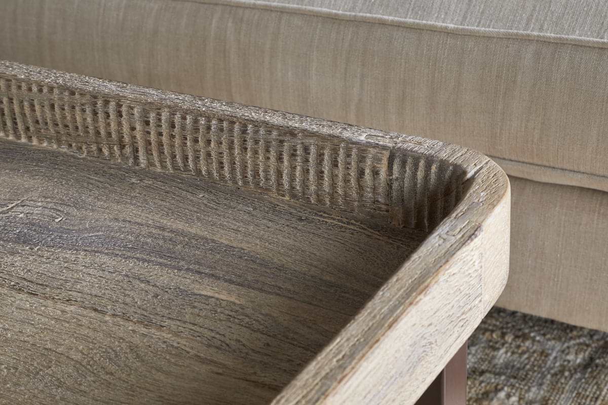 Close-up of the nkuku Nisha Reclaimed Wood & Iron Coffee Table - Natural, featuring a raised textured edge and weathered finish with visible wood grain. A beige upholstered sofa and part of a patterned rug appear in the background.