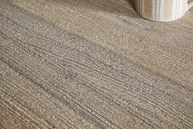 Close-up of the nkuku Nisha Reclaimed Wood & Iron Coffee Table - Natural, showing textured light brown wood with visible grain. In the upper right, part of a ceramic mug with beige and brown vertical stripes is seen.