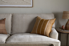 A beige fabric sofa is styled with nkuku’s Eesha Jute Cushion Cover in ochre. Against a light wall, it pairs with a striped brown cushion. A round wooden side table holds a textured lamp and candle beneath minimalist wall art.