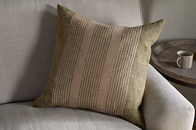The nkuku Eesha Jute Cushion Cover in sage green rests against a light gray sofa, adding modern rustic charm. A round wooden side table with a beige mug nearby enhances the cozy, inviting atmosphere.