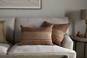 A cozy beige sofa is styled with modern rustic touches, including nkuku’s Eesha Jute Cushion Cover in Rust, brown striped and green pillows, a round wooden table with a lamp and candle, and textured wall art above in neutral tones.