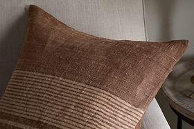 The Eesha Jute Cushion Cover - Rust by nkuku, featuring a brown textured finish with subtle beige stripes, rests on a light beige chair. Its modern rustic design adds warmth, complemented by a small round wooden side table in the background.