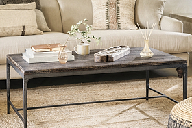 A cozy living room featuring the nkuku Sita Reclaimed Wood & Iron Coffee Table in Chocolate Brown, styled with books, a vase of greenery, a mug, wooden tray, and reed diffuser on a woven rug before a beige sofa with neutral-patterned pillows.