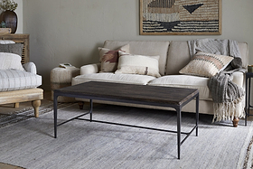 A cozy living room with a beige sofa, textured pillows, and a gray throw blends warmth and style. The nkuku Sita Reclaimed Wood & Iron Coffee Table in Chocolate Brown anchors the space atop a light woven rug, with baskets and wall decor for accents.