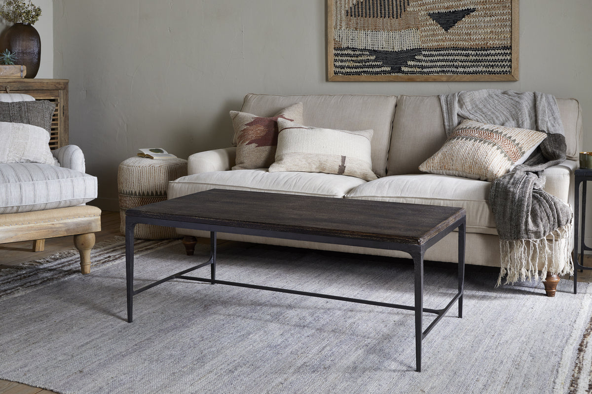 A cozy living room with a beige sofa, textured pillows, and a gray throw blends warmth and style. The nkuku Sita Reclaimed Wood & Iron Coffee Table in Chocolate Brown anchors the space atop a light woven rug, with baskets and wall decor for accents.