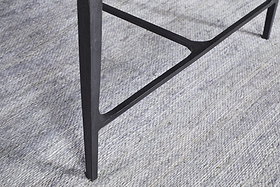 A close-up of a slim, dark leg from the nkuku Sita Reclaimed Wood & Iron Coffee Table in Chocolate Brown, with a triangular support bar, stands on a textured light gray carpet with subtle horizontal lines.