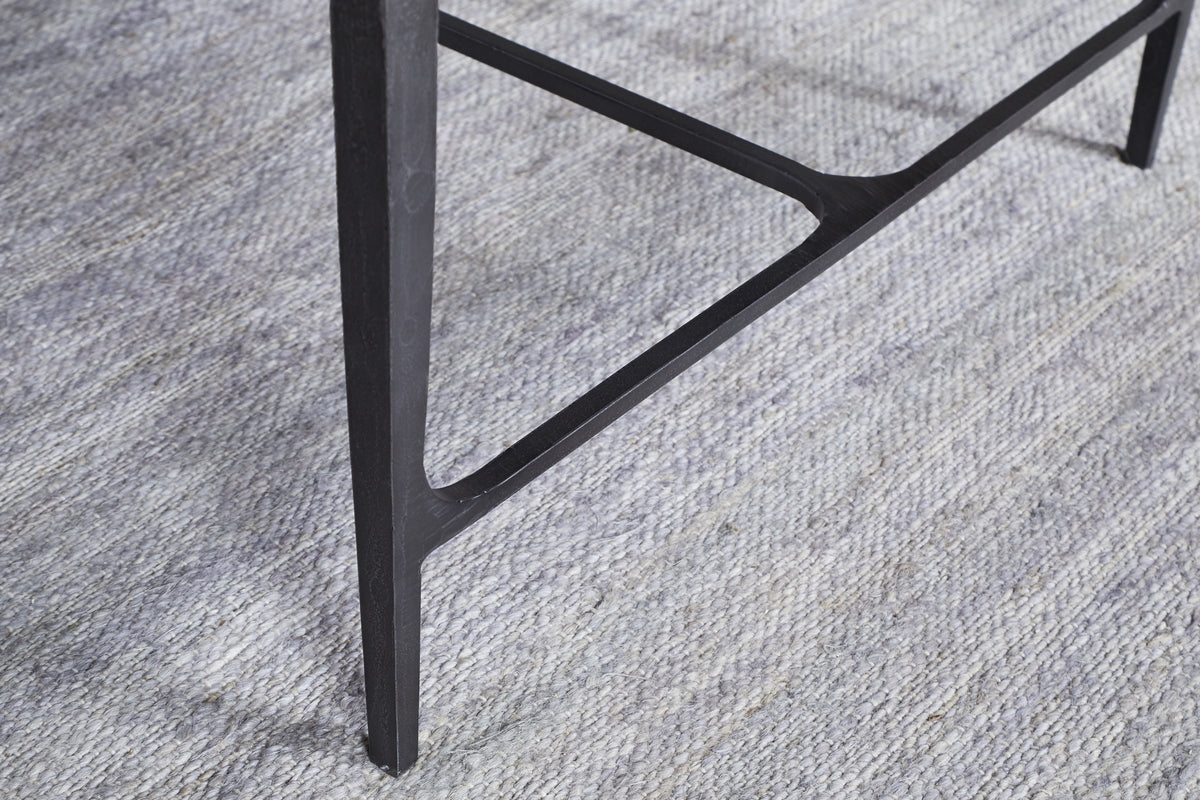 A close-up of a slim, dark leg from the nkuku Sita Reclaimed Wood & Iron Coffee Table in Chocolate Brown, with a triangular support bar, stands on a textured light gray carpet with subtle horizontal lines.