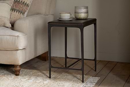The nkuku Sita Reclaimed Wood & Iron Side Table in chocolate brown stands by a beige sofa topped with a ceramic teacup, saucer, and glass jar candle. A woven rug peeks out beneath, while textured cushions accent the sofa.