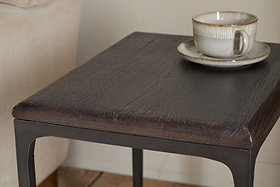 A close-up of the nkuku Sita Reclaimed Wood & Iron Side Table in chocolate brown features thin metal legs. A beige ceramic cup and saucer rest on top, with a beige sofa in the softly lit background—an inviting accent piece.