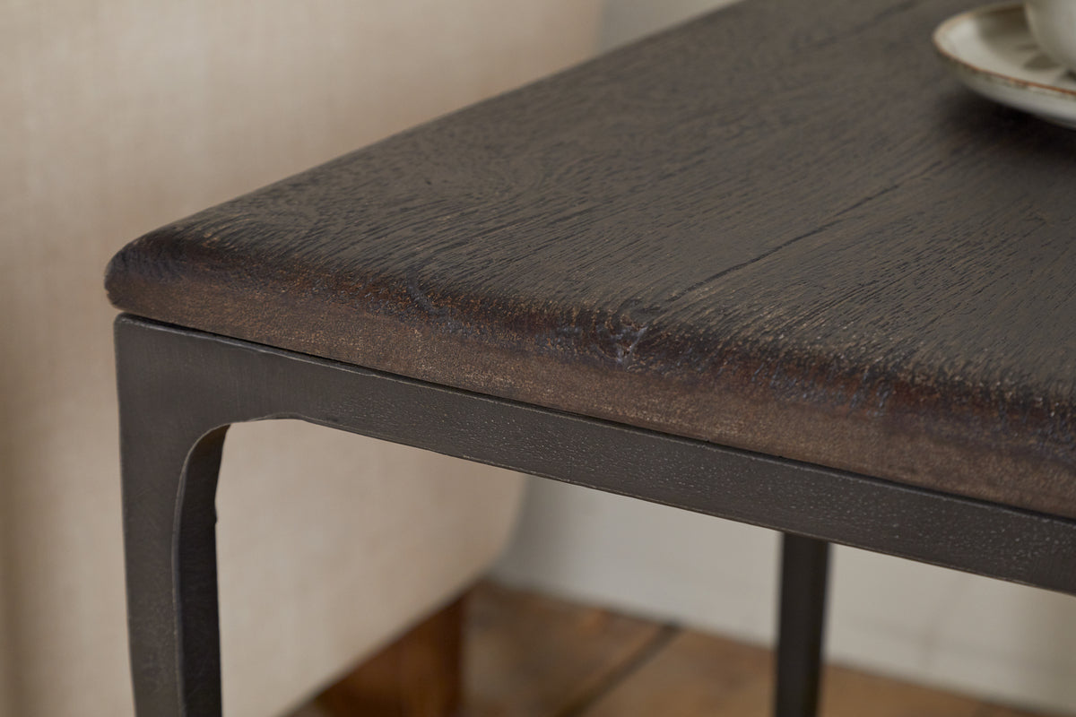 A close-up of the Sita Reclaimed Wood & Iron Side Table in Chocolate Brown by nkuku shows its smooth, rounded edge and black metal frame. A beige cushioned chair and ceramic plate are partly visible on a wooden floor in the background.
