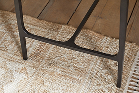 Close-up of the Sita Reclaimed Wood & Iron Side Table by nkuku, featuring a black iron leg resting on a textured beige woven rug. The rug overlays a chocolate brown reclaimed wood floor with visible grain and plank gaps.