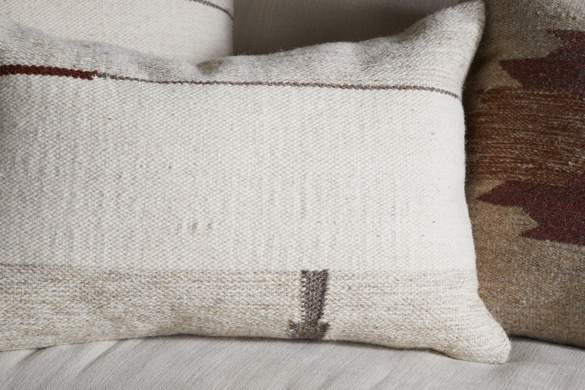 A close-up of two decorative pillows on a light fabric surface. The front showcases nkuku’s Tamil Wool Cushion Cover - Natural with an off-white woven texture and subtle brown lines; the back pillow displays a warm, earthy leaf pattern.