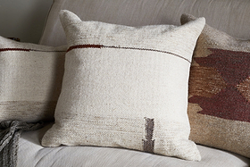 Three decorative cushions rest on a light beige sofa. The central cushion features the nkuku Tamil Wool Cushion Cover - Natural, with a minimal brown and dark red design, while charcoal and rust pillows add a cozy, rustic touch.