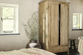 The nkuku Vivan Grooved Wardrobe - Natural with two doors stands by a big brown ceramic vase with tall branches. Cream walls, a beige knit blanket, black-framed windows, and a wooden stool complete the cozy bedroom storage setup.