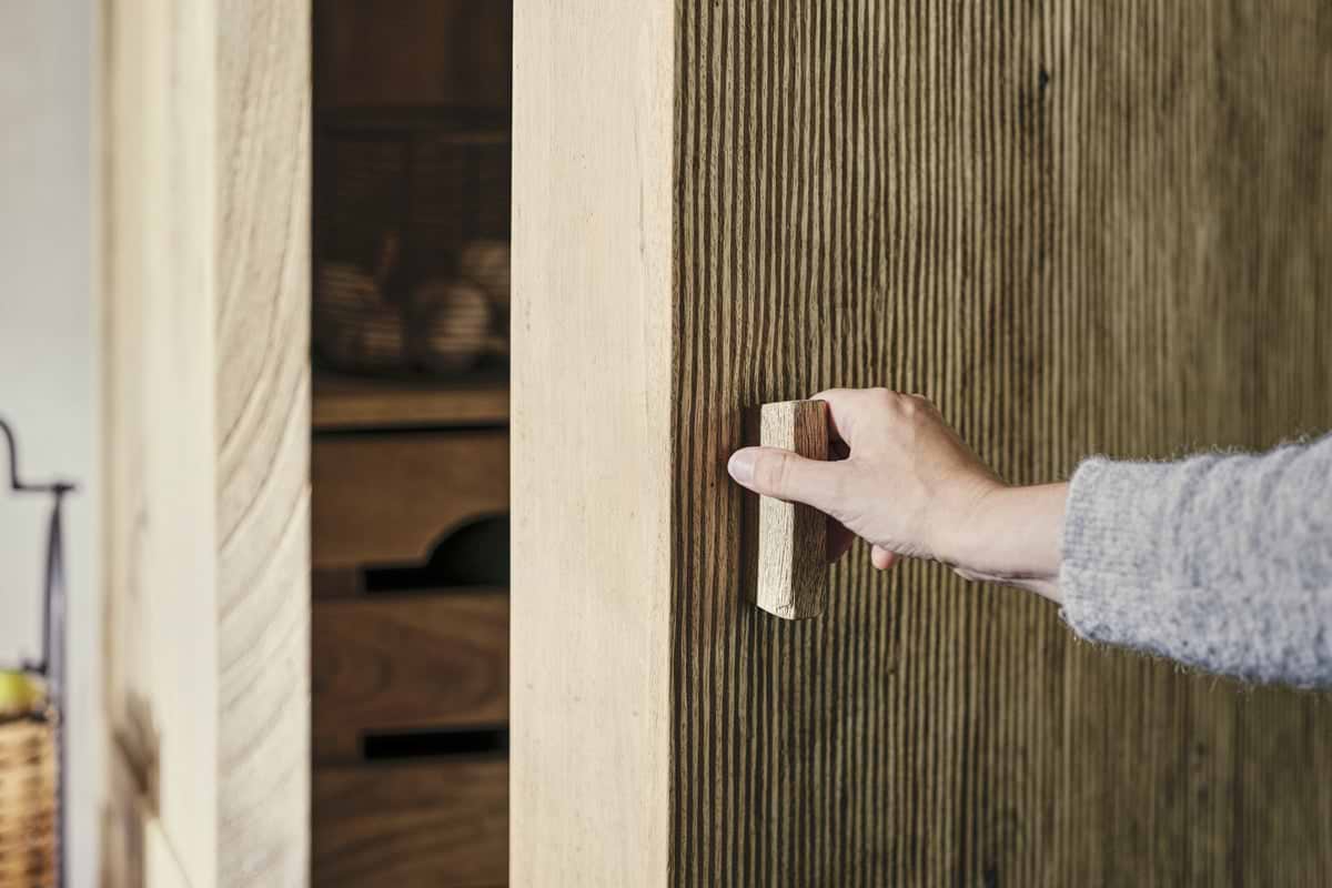 A person opens the nkuku Vivan Grooved Pantry Cupboard - Natural, featuring a textured ribbed wood surface and vertical handle. Inside the stylish wooden cabinet, shelves display baskets and storage boxes in the background.