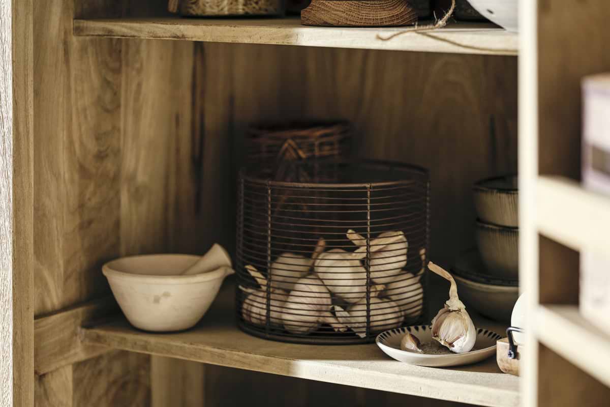 The nkuku Vivan Grooved Pantry Cupboard in natural wood brings charm to your kitchen, neatly storing a mortar and pestle, garlic bulbs in a wire basket, stacked ceramic bowls, and a plate of garlic cloves for a cozy, organized look.