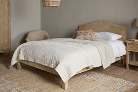 A cozy bedroom featuring the nkuku Vivan Grooved Bed in natural mango wood, complemented by a beige quilt, white sheets, decorative pillows, a matching wooden nightstand and wardrobe, woven chair, ceiling lamp, and a textured neutral rug on a wood floor.