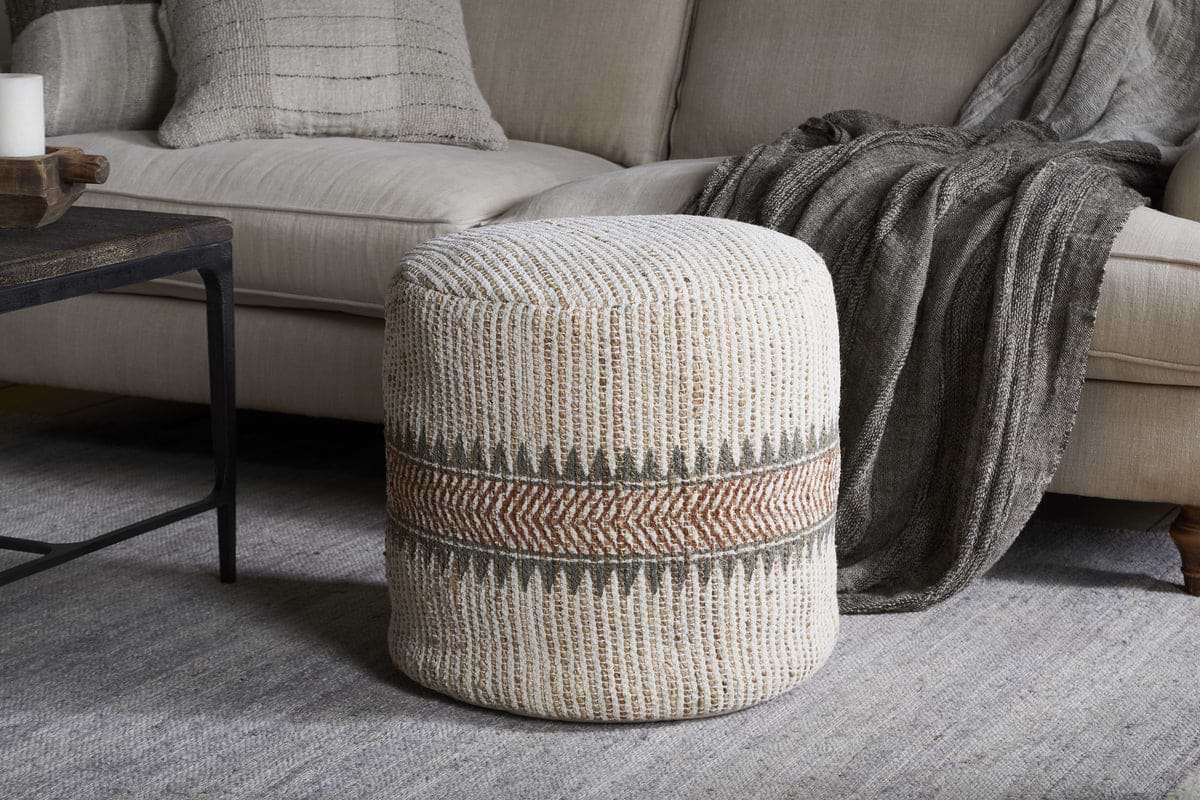 The nkuku Zairya Jute & Cotton Tall Pouf in natural and rust, featuring a textured cylindrical design with beige, green, and rust zigzag stripes, sits on a light gray rug before a beige sofa with gray accents and a dark wood coffee table.