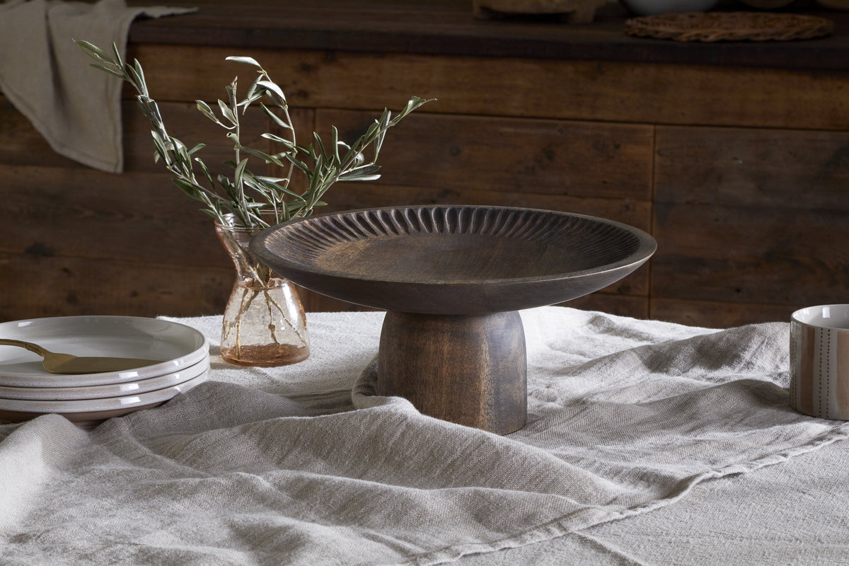 The Eisha Wooden Cake Stand – Brown by nkuku rests on a beige linen tablecloth, beside stacked white plates, a gold spoon, serveware essentials, a striped mug, and a glass jar with olive branches. A wooden cabinet is blurred in the background.
