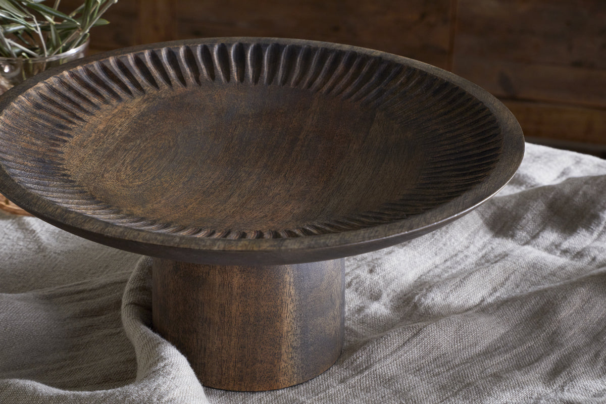 The Eisha Wooden Cake Stand by nkuku features a wide, ridged edge and cylindrical base, hand-carved from dark brown wood. Set on a light cloth, its rustic design is complemented by a blurred branch in the warm background.