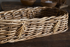 The Bersama Rattan Bread Basket - Natural by nkuku is an empty, oval handwoven basket made from light and dark brown rattan reeds. It sits on a rustic wooden surface with blurred wooden bowls adding charm to the warm tabletop decor.